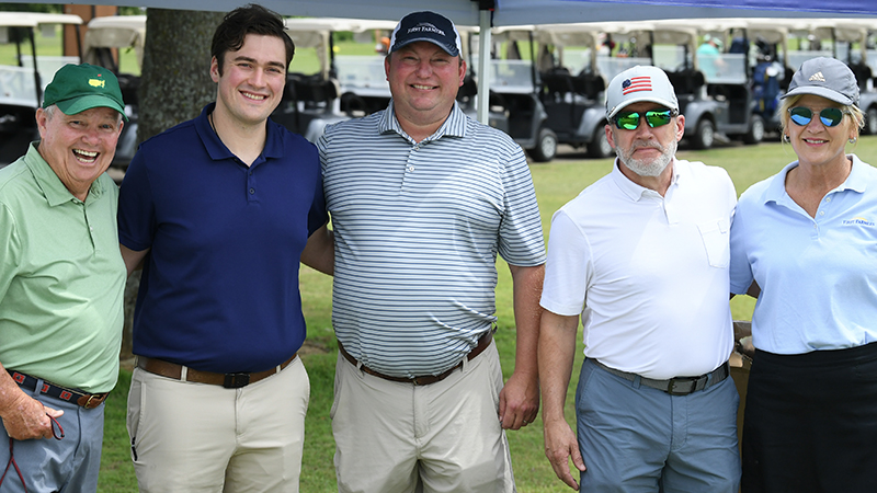 Five golfers stand together outdoors on a golf course, with golf carts in the background.
